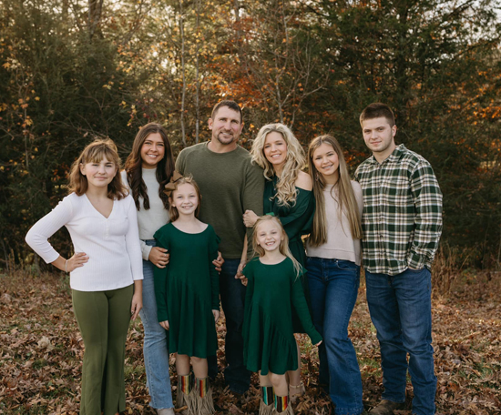 A family posing outdoors in a wooded area during autumn, dressed in coordinated green and neutral outfits, smiling at the camera.