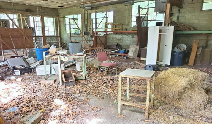 A cluttered indoor space filled with old furniture, scrap materials, hay bales, and scattered debris, with shelves and windows in the background.